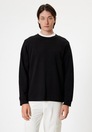 Sudadera negra de manga larga hecha de tela suave, con cuello redondo y corte relajado. Llevada sobre una camiseta blanca. Fondo blanco.