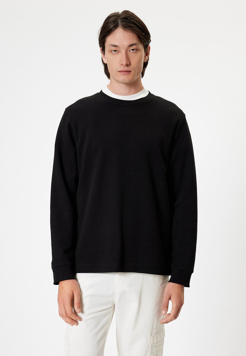 Svart långärmad sweatshirt tillverkad av mjukt tyg, med rund hals och avslappnad passform. Bärs över en vit undertröja. Vit bakgrund.