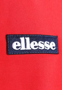 Rødt stof med en rektangulær marineblå lap med hvidt broderet tekst "ellesse" i en fed skrifttype.