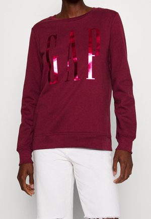 Person iført en bordeauxrød sweatshirt med lange ærmer og en skinnende rød "GAP"-logo samt hvide bukser, stående mod en ensfarvet hvid baggrund.