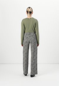 Donna con capelli biondi raccolti in una coda di cavallo, indossa un top a maniche lunghe verde oliva e pantaloni larghi grigio a fantasia zebrate, in piedi di fronte a un muro bianco semplice.