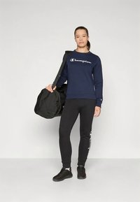 Nuori nainen seisoo urheiluvaatteissa, pitäen mustaa urheilulaukkua olkansa päällä vaaleaa taustaa vasten.