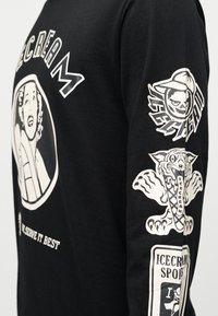 Μαύρο μακρυμάνικο πουκάμισο με λευκές γραφικές εκτυπώσεις. Το σχέδιο περιλαμβάνει το πρόσωπο μιας γυναίκας, μια αρκούδα με καπέλο και μια τίγρη που κρατάει έναν χωνάκι παγωτού.