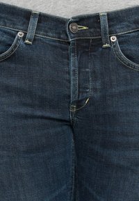 Gros plan de la ceinture d'un jean en denim bleu foncé avec bouton métallique et poches avant sur un fond de chemise gris clair.