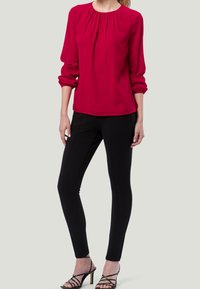 Blouse rouge avec un col froncé et des manches longues, portée avec un pantalon noir slim. L'ensemble est complété par des talons à lanières.
