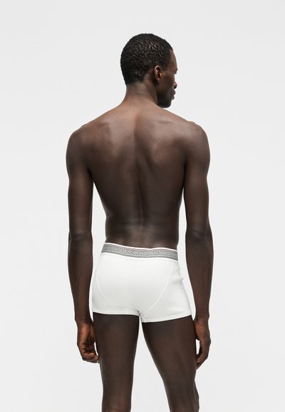 Modèle masculin vu de dos, portant un boxer blanc avec une ceinture noire à motifs, sur un fond gris clair uni.