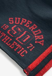 Marineblaues Gewebe mit rotem Drucktext. Enthält "SUPERDRY" und "ATHLETIC" mit strukturierten Details. Beinhaltet einen roten Akzent am Saum.