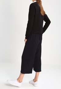 Donna che indossa un top nero a maniche lunghe e pantaloni neri larghi tagliati sopra la caviglia, con scarpe da ginnastica bianche, in piedi con una mano sul fianco.