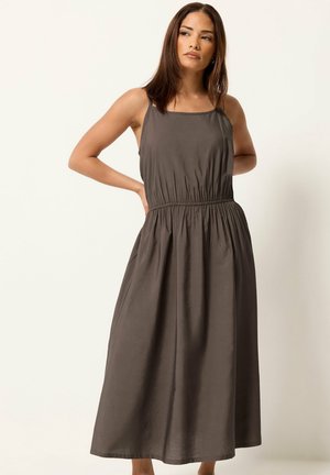 Femme en robe midi gris foncé sans manches avec taille froncée, debout les mains derrière le dos, regardant sur le côté sur fond uni.
