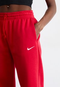 Červené Nike teplákové nohavice s elastickým pásom a šnúrkou. Obsahujú bočné vrecká a malé biele logo na ľavom stehne.