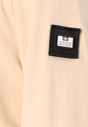 Tissu beige au toucher doux, comportant un patch carré noir avec l'inscription "WEEKEND OFFENDER" et un logo blanc.