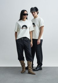 Deux mannequins portent des tenues coordonnées : des t-shirts crème oversize avec un imprimé graphique, des pantalons noirs à motifs, et des bottes texturées.