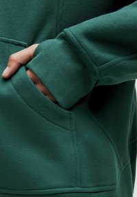 Felpa verde realizzata in un tessuto morbido, con tasca a marsupio e polsini a coste. La texture appare liscia con una leggera lucentezza.