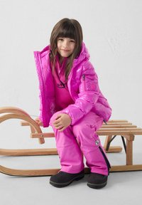 Roze opblaasbare jas met capuchon, gecombineerd met bijpassende roze skibroek. Zwarte waterdichte laarzen en een houten slee op de achtergrond.