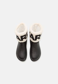 Botas de cuero negras con un puño de forro polar texturizado blanco que presenta letras en negrita en negro. Exterior suave con puntas redondeadas y suelas planas.