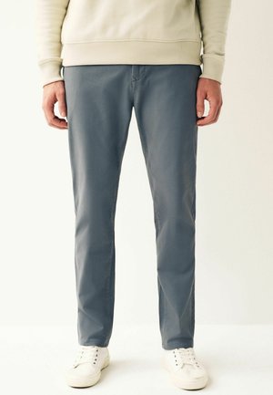 Grey cotton trousers with a straight cut, front pockets, and a slight sheen. Paired with white sneakers for a casual look.