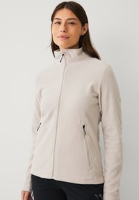 Giacca in felpa beige chiaro con chiusura a zip, colletto alto e due tasche laterali con zip. Tessuto morbido e strutturato con un design aderente.