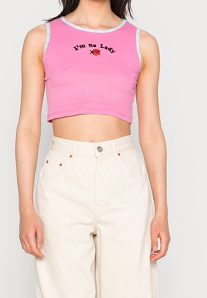 Kobieta ubrana w różowy bezrękawnik typu crop top z napisem „I'm no Lady” oraz grafiką biedronki, zestawiony z kremowymi spodniami z wysokim stanem.