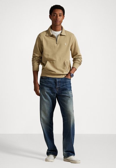 Beige ritssluiting pullover met een voorzak, gecombineerd met vervaagde blauwe jeans en witte sneakers. Accessoire bestaat uit een zilveren horloge.
