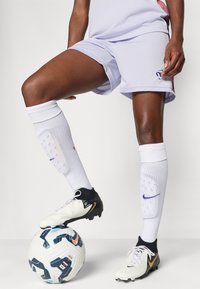 Weiße Fußballshorts mit lila Akzenten, kombiniert mit weißen kniehohen Socken und schwarz-weißen Stiefeln, abgelegt auf einem strukturierten Fußball.