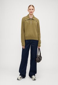 Suéter tricotado verde-oliva com um colarinho largo e fecho de meia-zíper, combinado com jeans de perna larga azul-escuro e uma pequena mala escura.