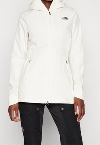 Giacca impermeabile bianca con chiusura a zip, cappuccio regolabile, tasche laterali e logo. Abbinata a pantaloni neri per un contrasto di stile.