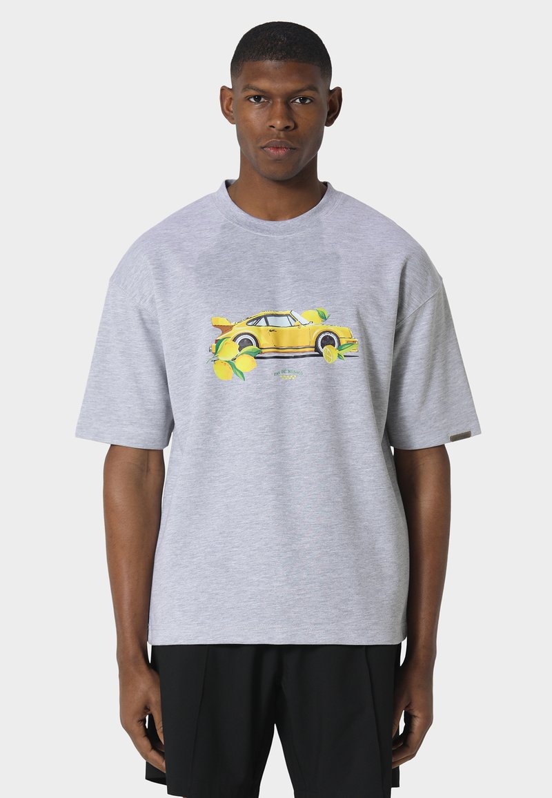 Homme portant une chemise à manches courtes gris clair avec un motif de voiture vintage jaune et des citrons, associée à un short noir, debout devant un fond uni.
