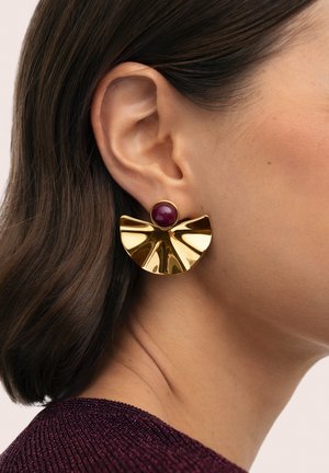 Oreja izquierda de una mujer que lleva un pendiente dorado en forma de abanico con una piedra redonda de color rojo oscuro, cabello marrón oscuro, y una blusa texturizada de color púrpura oscuro.