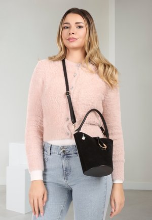 Jeune femme portant un cardigan rose pelucheux et un jean bleu clair, tenant un sac à main noir avec une bandoulière portée en travers du corps.