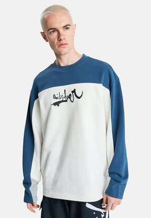 Młody mężczyzna z krótkimi blond włosami, ubrany w niebiesko-białą koszulkę z długim rękawem z czarnym logo w stylu pisma na piersi, stoi i patrzy w lewo.