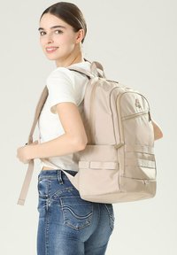 Sac à dos en tissu beige avec une poche avant zippée, des bretelles réglables et des poches latérales. Il présente un design épuré et une surface texturée.