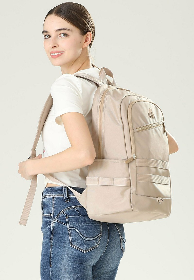 Sac à dos en tissu beige avec une poche avant zippée, des bretelles réglables et des poches latérales. Il présente un design épuré et une surface texturée.