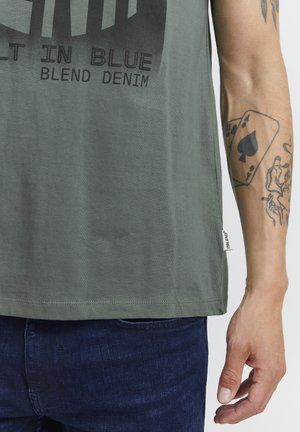 Olīvzaļš T-krekls ar drukātu tumšu grafiku un tekstu. Kokvilnas maisījuma audums ar īsām piedurknēm un brīvu piegriezumu. Iespējama zīmola etiķete.