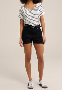 Frau trägt ein weißes und schwarzes gestreiftes V-Ausschnitt-T-Shirt, schwarze hochgeschnittene Shorts mit Gürtel und weiße Sneakers mit schwarzen Streifen.