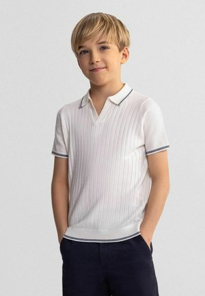 Garçon blond, mains dans les poches, portant un polo en maille blanc à manches courtes avec une fine bordure noire et un pantalon foncé, debout devant un fond gris uni.