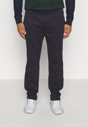 Pantalones de chándal azul marino oscuro con una textura suave, ajuste ceñido, cinturilla elástica y bolsillos laterales, combinados con zapatillas deportivas blancas.