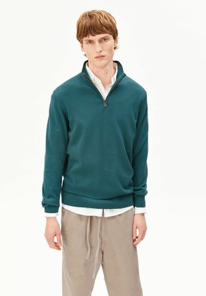 Junger Mann trägt einen türkisfarbenen Pullover mit Viertelreißverschluss über einem weißen Hemd und beigen Cordhosen, steht vor einem schlichten weißen Hintergrund.