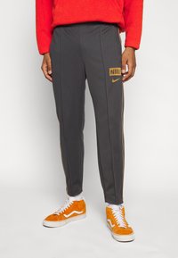 Grå sportbyxor med en gyllene Nike-logotyp på vänster lår, har guld sidostråkar och en smal passform, tillsammans med orange sneakers.