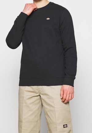 Osoba nosząca czarną bluzę z długim rękawem i beżowe spodnie, oba z małymi prostokątnymi logo Dickies na piersi i nodze.