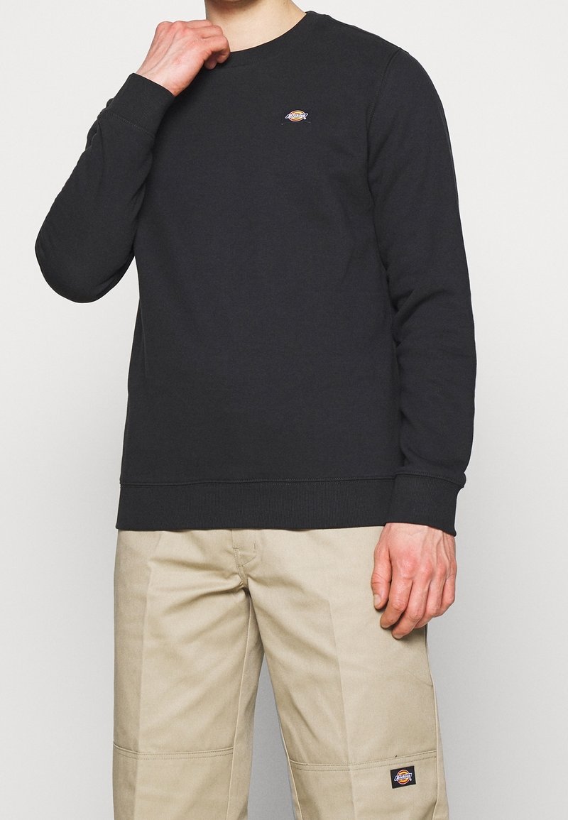 Personne portant un sweat-shirt noir à manches longues et un pantalon beige, tous deux avec de petits logos rectangulaires Dickies sur la poitrine et la jambe.