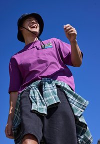 Jeune homme riant portant un t-shirt violet, un chapeau bob noir, un short noir et une chemise à carreaux verte nouée autour de la taille devant un ciel bleu clair.