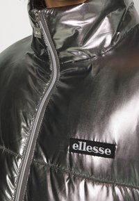 Metalická stříbrná prošívaná bunda s vysokým límcem, předním zipem a vyšívaným černým logem "ellesse" na hrudi. Hladká textura.