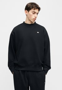 Νεαρός άνδρας που φοράει μαύρο φούτερ και μαύρο παντελόνι, κοιτάζοντας προς τα κάτω αριστερά με τα χέρια στις τσέπες, σε απλό ανοιχτό φόντο.