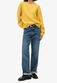 Maglione giallo, lavorato a maglia, con scollo rotondo e spalle scese, abbinato a jeans azzurri chiari, morbidi e arrotolati ai polsi, e scarpe nere.