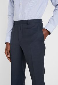 Marineblaue Anzughose aus einem strukturierten Stoff, mit Seitentaschen und geradem Bein, kombiniert mit einem hellblauen gemusterten Hemd.