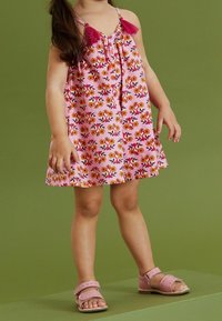Robe fleurie rose avec des motifs multicolores et des franges roses. Associée à des sandales rose clair avec une bordure festonnée.