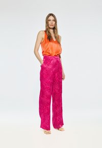 Feloranje satijnen tanktop gecombineerd met losse roze broek met een gestructureerd dierenprintontwerp. Model draagt gouden sandalen.