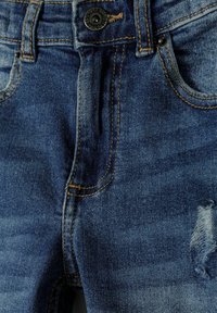 Vaqueros de mezclilla en azul oscuro con una textura ligeramente desgastada. Presenta un solo botón de metal y costuras decorativas, incluyendo detalles desgastados.
