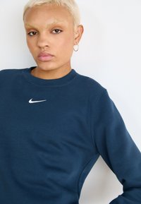 M�ørkeblå genser laget av mykt stoff, med rund hals og en liten hvit Nike-logo på brystet.