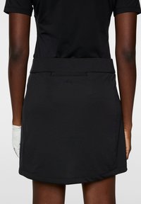 Svart golfkjol med en slät textur, som har en platt midja och en bakficka. En liten logotyp syns nära sömmen på midjan.